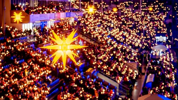 Ein Herrnhuter Stern hängt über einer Tribüne voller Menschen mit Kerzen