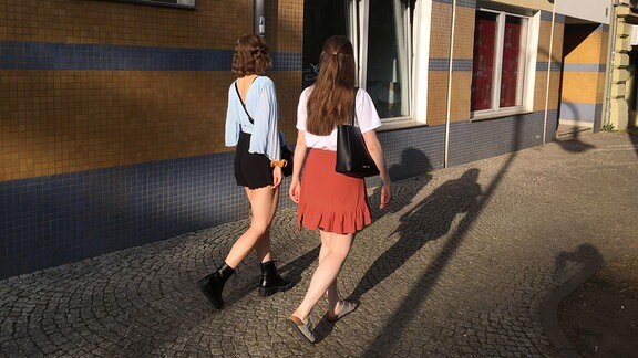  Zwei junge Frauen in Sommerkleidung auf dem Fußgängerweg sind von hinten zu sehen.