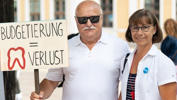 Ein Mann und eine Frau blicken in die Kamera, neben ihnen ein Schild mit der Aufschrift "Budgetierung gleich Zahnverlust"