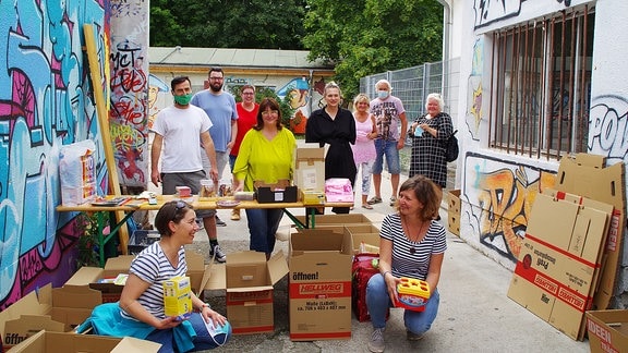 Eine Gruppe von Helfer:innen steht und kniet im Innenraum eines Innenhofes und präsentiert fröhlich Kisten mit Spenden. 