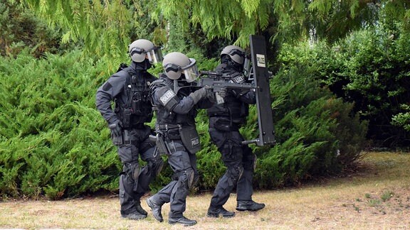 Drei Polizisten in Spezialausrüstung schleichen sich mit Schutzschild und Maschinengewehr an.
