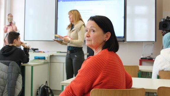Ältere schwarzhaarige Frau in rotem Pullover sitzt in einem Klassenraum und blickt über die Schulter