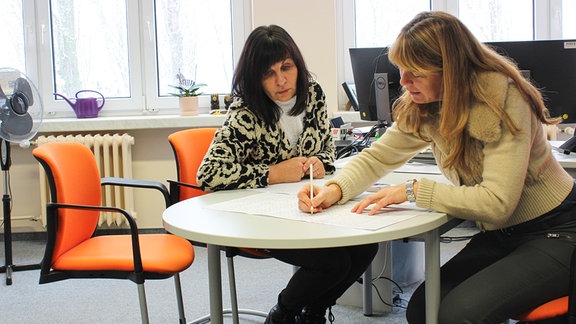 Zwei Frauen sitzen an einem runden Tisch in einem Büro über Plänen auf großen Papieren