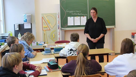 Eine Frau mit dunkelblonden langen Haaren und schwarzer Kleidung unterrichtet in einem Klassenzimmer mit größeren Kindern