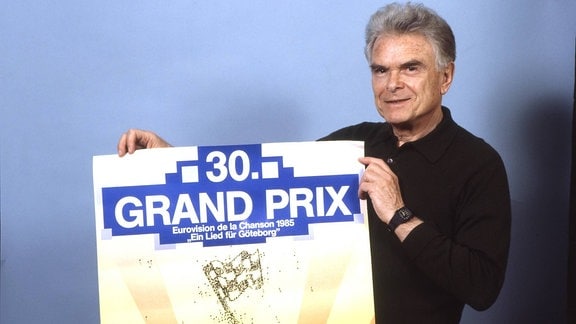 Ein grauhaariger Mannn hält ein Plakat vom Grand Prix Eurovision de la chanson 1985 hoch.
