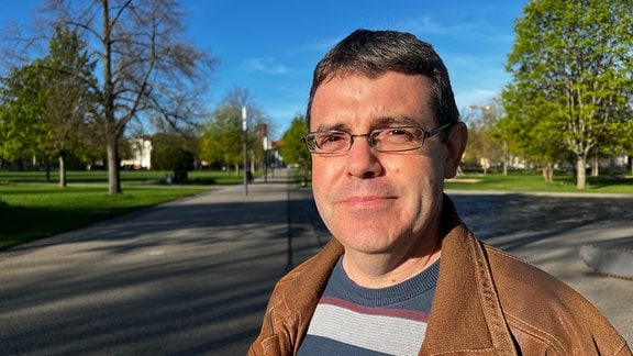 Ein Mann mit Brille, kurzen schwarzen Haaren und brauner Jacke blickt in die Kamera. Im Hintergrund ist eine Parkanlage zu sehen.