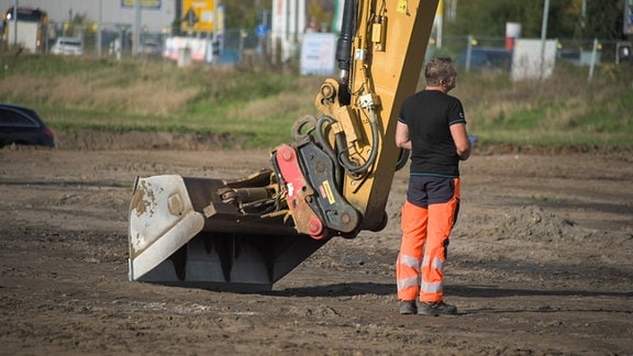 Auf einer Freifläche in einem Gewergegebiet in Schönebeck steht ein Mann in Warnkleidung vor einer Baggerschaufel.