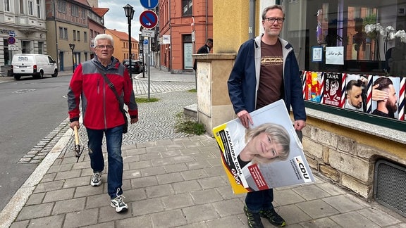 Zwei Männer mit laufen mit einem Wahlplakat in der Hand durch die Stadt.