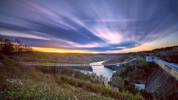 Eine lange Seilhängebrücke führt über einen Fluss, der Himmel schimmert bläulich, die Sonne geht unter.  