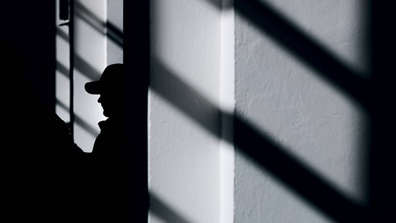 Symbolbild: Die Silhouette eines Mannes mit Basecap zeichnet sich zwischen Schatten eines Fensters ab.