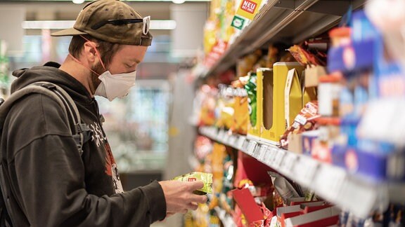 Ein Kunde steht in einem Supermarkt vor einem Regal, und trägt dabei einen Mundschutz. 