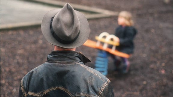 Ein Mann beobachtet ein spielendes Kind auf einem leeren Spielplatz.