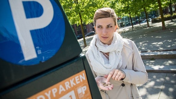 Eine junge Frau vor einem Parkscheinautomat hält Münzen in ihrer Hand. (Inszeniertes Foto)