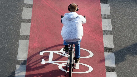 Ein Fahrrad ist auf einem neuen, roten Radweg auf der Schlossstrasse in Steglitz aufgemalt. Ein junger Mann auf einem Rad, der bei der Fahrt Kopfhoerer traegt, faehrt auf dem Radweg.