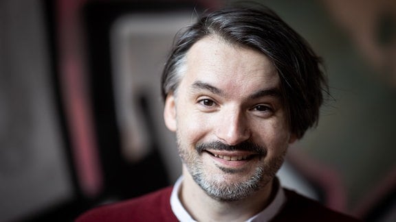 Porträt Saša Stanišić: Ein Mann mit Pullover und Hemdkragen lächelt breit in die Kamera.