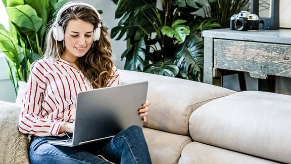 Eine ächelnde Frau mit Laptop und Kopfhörern sitzt auf einer Couch