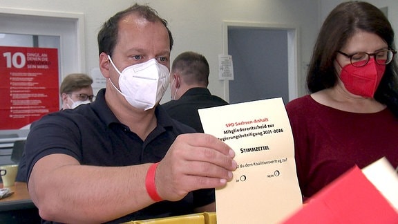 Ein Mann mit Mund-Nasen-Schutz hält einen Stimmzettel hoch.