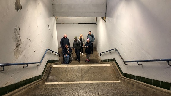 Eine Gruppe Menschen, einer davon im Rollstuhl, steht vor einem Treppenaufgang am Bahnhof