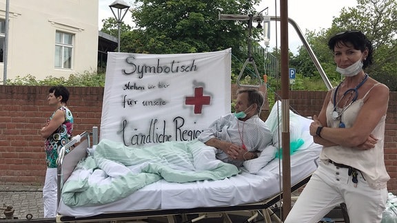 Ein gestellter Wagen mit einem Mann im Krankenbettt und einer gestellten Krankschwester vor ihm sowie der Aufschrift "Symbolisch stehen wir hier für unsere ländliche Region" auf einem Banner.