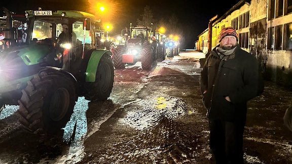 Ein Mann steht nachts neben einem Traktoren-Konvoi.