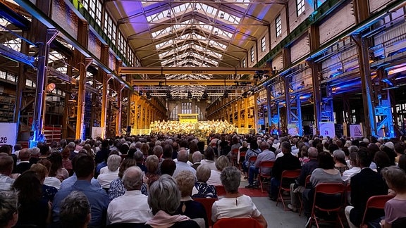 Eine Bühne in einem Industriegebäude mit voll besetzten Stühlen im Publikum.