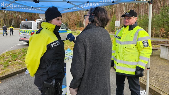 Eine Radioreporterin befragt mit dem Mikrofon zwei Polizisten in auffälliger neongelber Kleidung