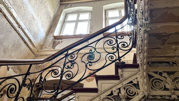 Blick nach oben auf ein verschörkeltes Treppengeländer vor alten Fenstern, von denen die Farbe abbröckelt