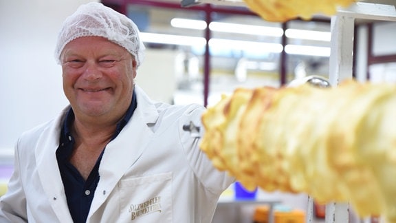 Der Geschäftsführer der Salzwedeler Baumkuchen GmbH, Frank Ostendorf, steht hinter einem Regal mit fertig gebackenen Baumkuchen.