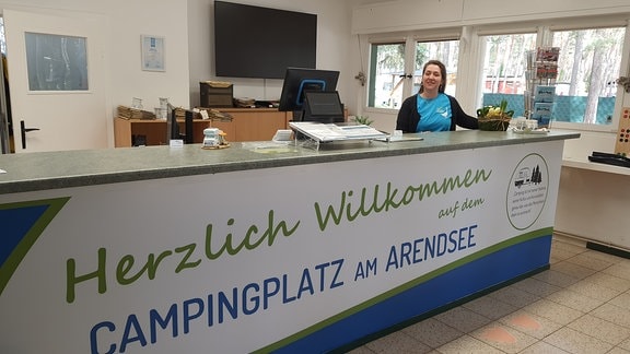 Frau hinter einem Tresen mit der Aufschrift "Campingplatz in Arendsee".