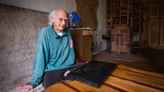 Ein alter Mann mit weißem Haarkranz und altem Pullover sitzt auf einer Bank vor einem Laptop und einem Ofen.
