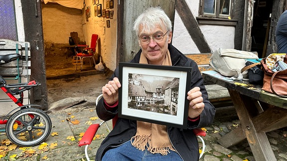 Ein Mann sitzt vor einem Fachwerkhaus auf einem Stuhl. Er zeigt ein gerahmtes Foto in die Kamera.
