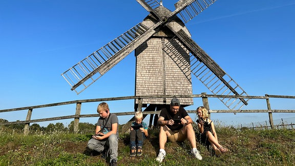 Eine Gruppe Erwachsene und Kinder mit Tablets pausieren während einer Museumstour vor eine historischen Windmühle.