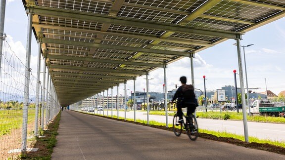 Ein Radfahrer fährt auf einem mit Solarmodulen überdachten Radweg entlang. 
