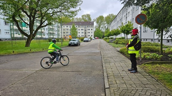 Ein junger Schüler mit Warnweste fährt auf einem Fahrrad vor an einem Mann mit Warnweste vorbei, der auf dem Bürgersteig steht.
