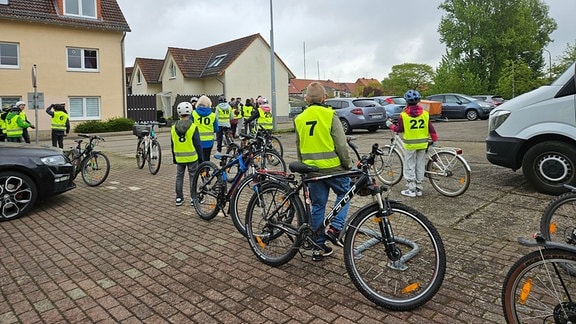 Zur Arbeit der Verkehrswacht gehört auch die Fahrradprüfung der vierten Klasse. Schüler:innen mit nummerierten Warnwesten und Fahrrädern stehen auf einem Hof.