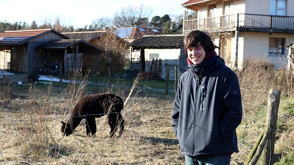 Ein junger Mann steht vor einer abgezäunten Wiese mit einem braunen Alpaka.
