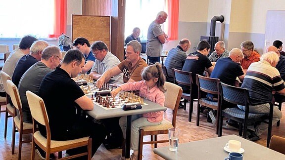 Ein braunhaariges Mädchen spielt mit zahlreichen Erwachsenen Schach.
