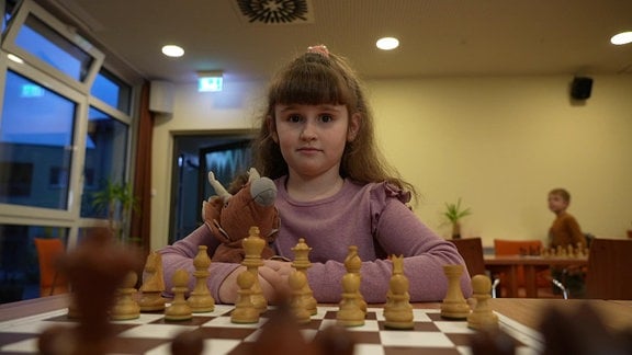 Ein braunhaariges Mädchen spielt Schach und hält einen Plüschdino.