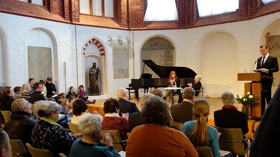 Zwei Frauen sitzen auf einem Podium in einer Kirche mit einem schwarzen Klavierflügel im Hintergrund. Ein Mann in Anzug spricht an einem Rednerpult. Im Vordergrund sind Menschen aus dem Publikum von hinten zu sehen.