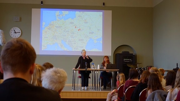 Zwei Frauen sitzen auf einem Podium in einer Kirche mit einer an die Wand gebeamten Karte Europas im Hintergrund. Im Vordergrund sind Menschen aus dem Publikum von hinten zu sehen.