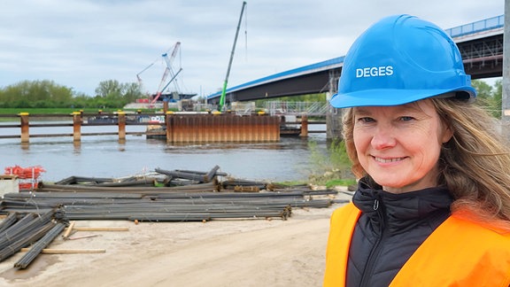 Frau in Warnweste und blauem Schutzhelm steht vor Baustelle an der Elbe