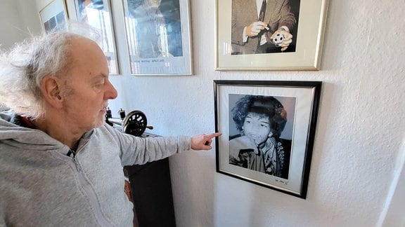 Ein Mann mit grauen Haaren steht vor einer Wand und zeigt auf ein dort hängendes Foto.