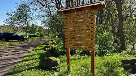 Eine hölzerne Hinweistafel des Natur- und Heimatvereins zeigt mit Pfeilen auf Sehenswertes in der Umgebung, wie Findlinge oder Bienenhaus