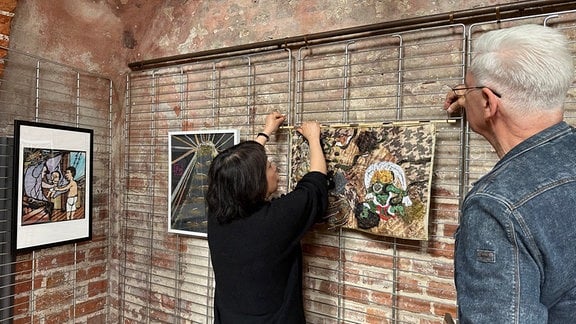Eine Frau hängt Bilder an einer Backsteinwand auf, ein Mann steht daneben.