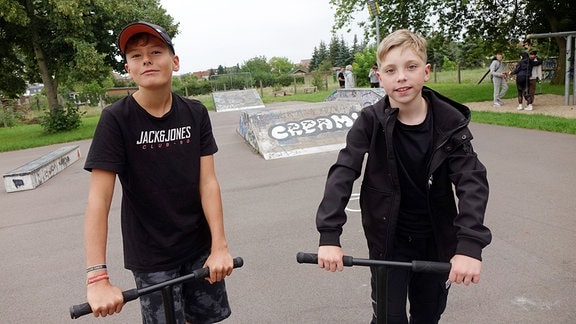 Zwei dunkelgekleidete Jungs auf Rollern auf einem Skateplatz.