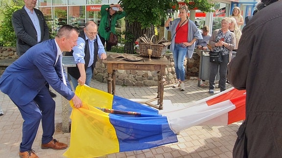 Mehrere Personen ziehen eine Flagge von einem zuvor verhüllten Spargeldenkmal. Das Denkmal zeigt einen Tisch auf dem ein Korb mit Spargel steht. 