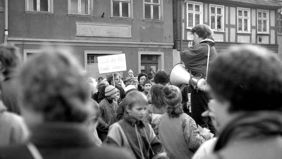 Historische Schwarzweiß-Aufnahme: Menschen demonstieren vor historischen Gebäuden. Im Vordergrund eine Frau mit Megafon.