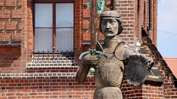 Steinfigur eines Ritters mit Schwert und Wappen vor einem Backsteingebäude 