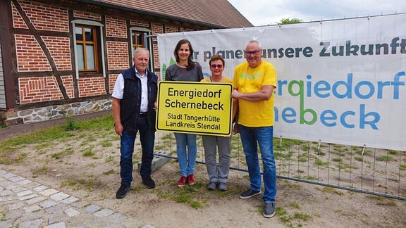 Vier Menschen stehen vor einem Fachwerkhaus und halten ein Orteingangsschild in der Hand mit der Aufschrift: Energiedorf Schernebeck.