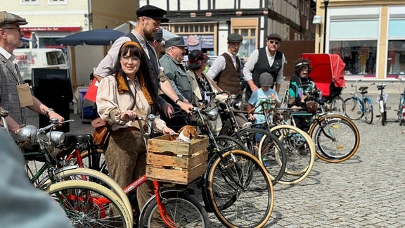 Einige Menschen stehen mit ihren Fahrrädern auf einem Marktplatz. Outfits und Rädern erinnern an die 1920er.